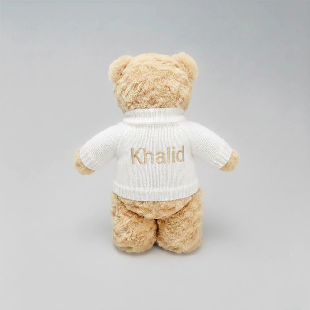 Beige teddy bear wearing a white sweater with 'Khalid' embroidered on a gray background