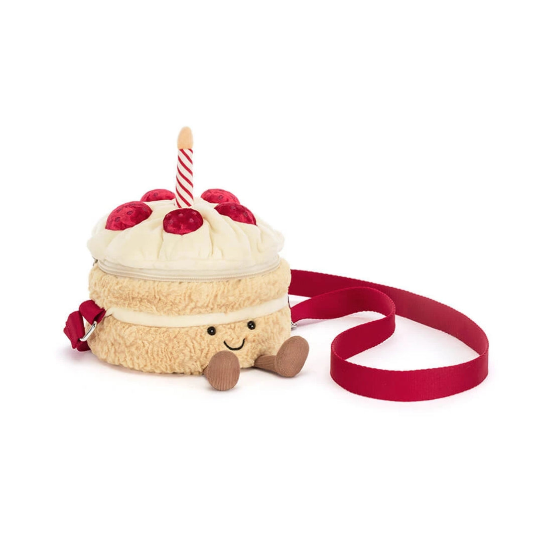 Birthday cake-shaped crossbody bag with cream frosting, red berry decorations, striped candle detail, smiling face, brown corduroy feet and a red shoulder strap.