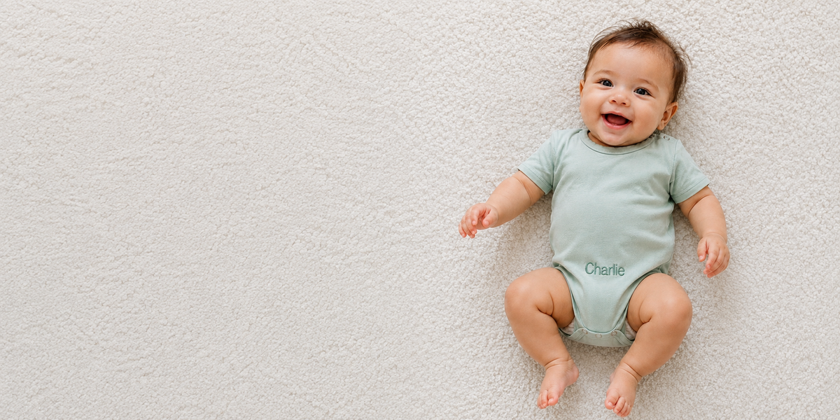 A baby lying on a textured white blanket, wearing a pale sage green short-sleeve bodysuit with the name 'Charlie' printed in dark green on the front.
