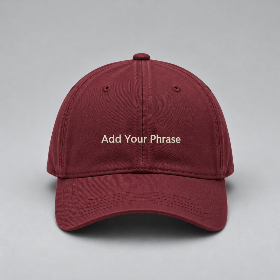Wine red baseball cap embroidered with “Add Your Phrase” in cream block lettering, featuring a curved brim and classic six-panel design against a light grey studio background.