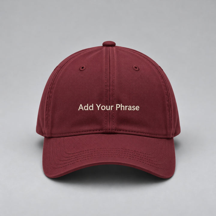 Wine red baseball cap embroidered with “Add Your Phrase” in cream block lettering, featuring a curved brim and classic six-panel design against a light grey studio background.