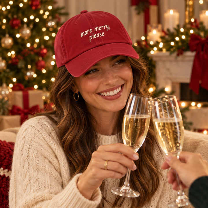 Woman wearing a red “more merry, please” embroidered Christmas cap, toasting champagne glasses beside a decorated Christmas tree.