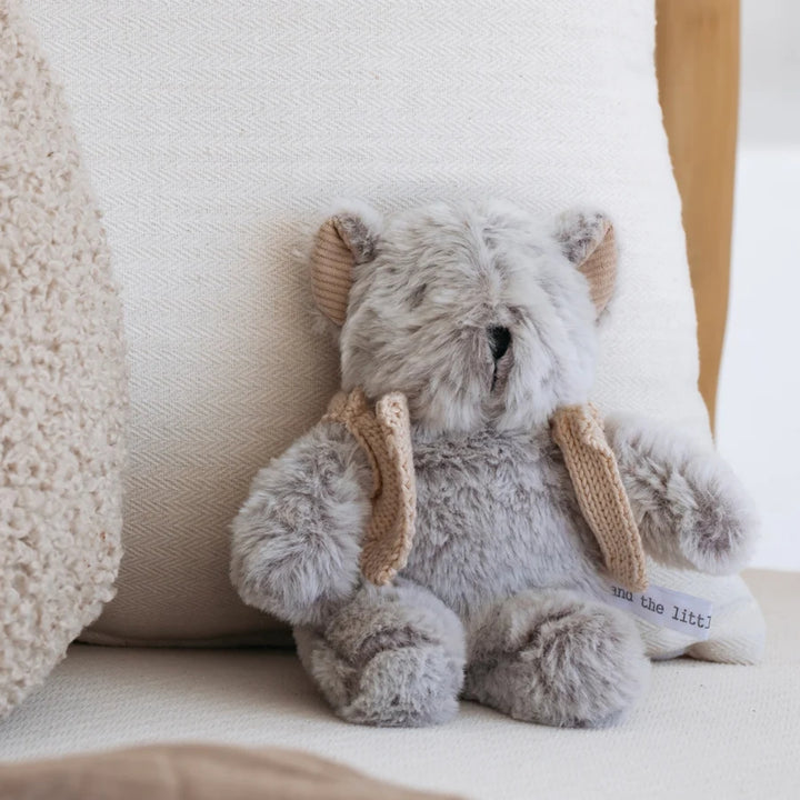 Soft grey teddy bear with beige knitted details sitting on a cream upholstered chair in a neutral home setting.