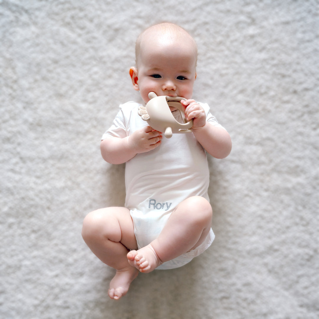 Baby wearing personalised white bamboo babygrow with "Rory" personalised in grey thread and holding Beige Chick Teether