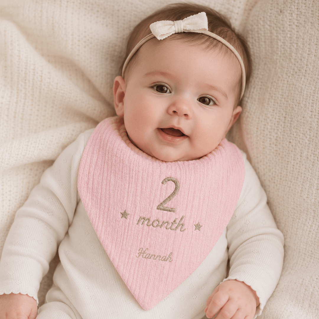 Baby wearing a pink bib with '2 month' and 'Hannah' text, sitting on a beige blanket.