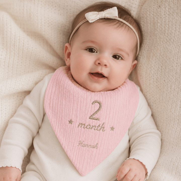 Baby wearing a pink bib with '2 month' and 'Hannah' text, sitting on a beige blanket.