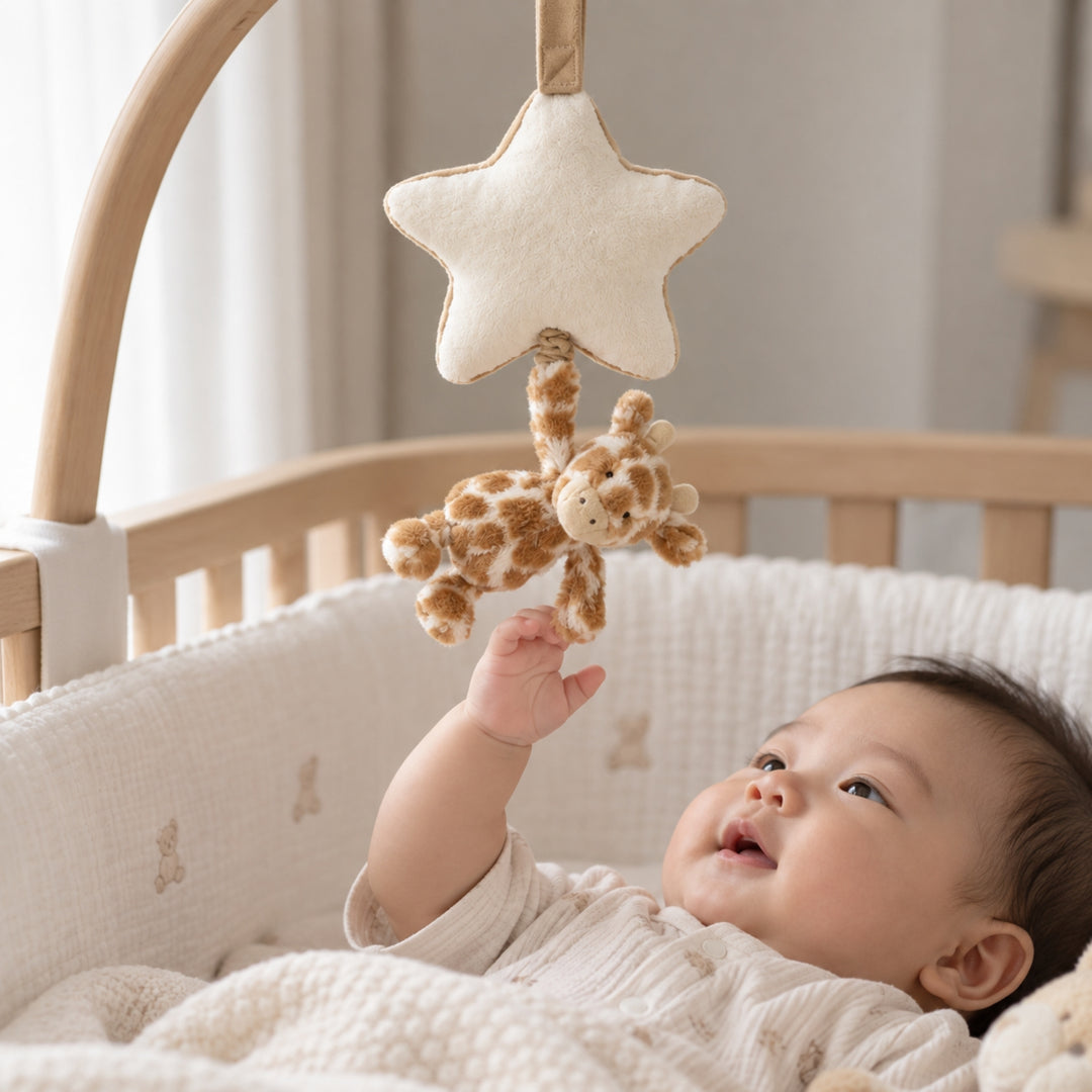 Baby lying in a wooden cot and reaching up towards a plush giraffe musical pull hanging from a cream star, photographed in a softly lit neutral nursery.