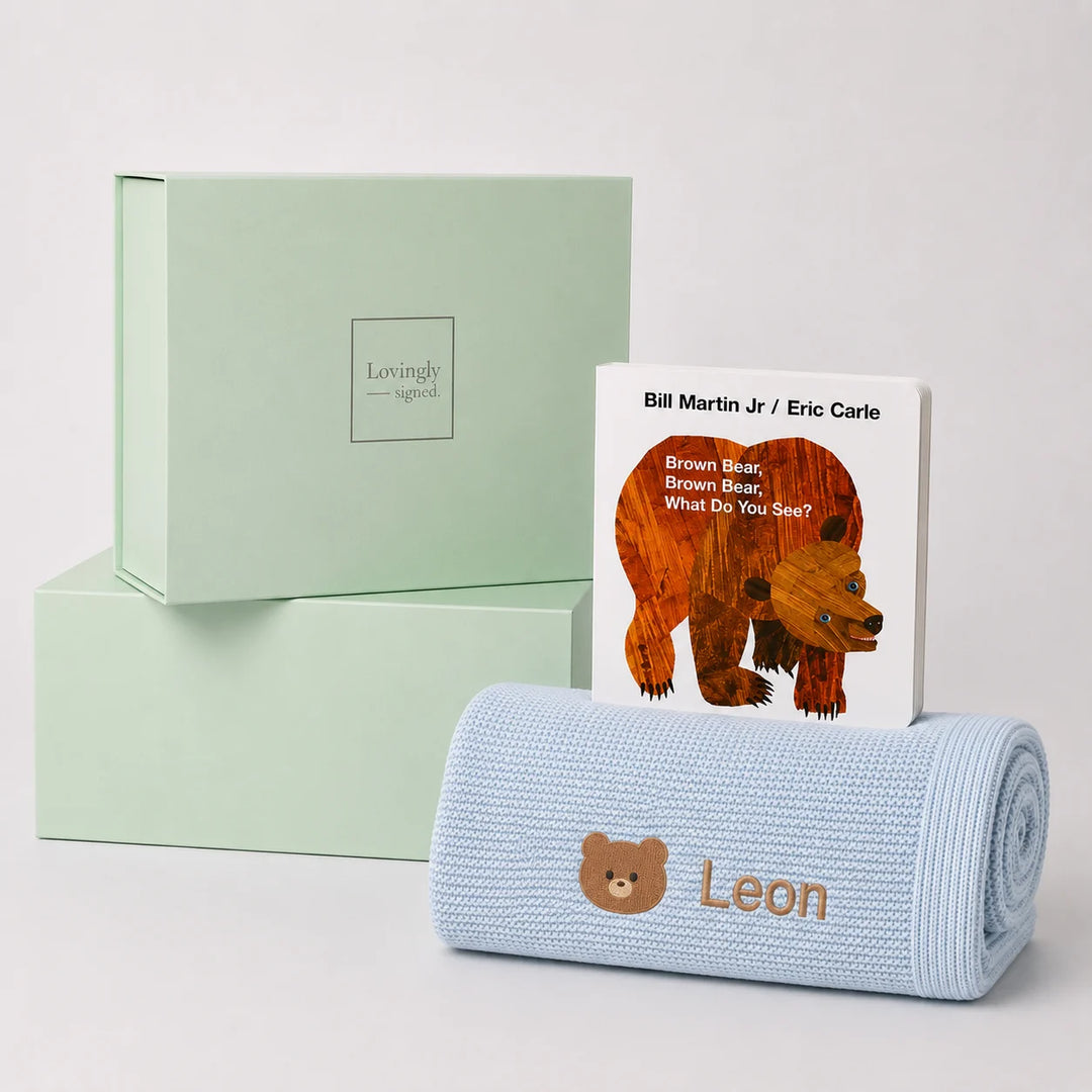 A personalized baby gift set featuring a light blue knit blanket with the name 'Leon' and a teddy bear face, a children's book titled 'Brown Bear, Brown Bear, What Do You See?' by Bill Martin Jr. and Eric Carle, and two mint green gift boxes from Lovingly Styled.