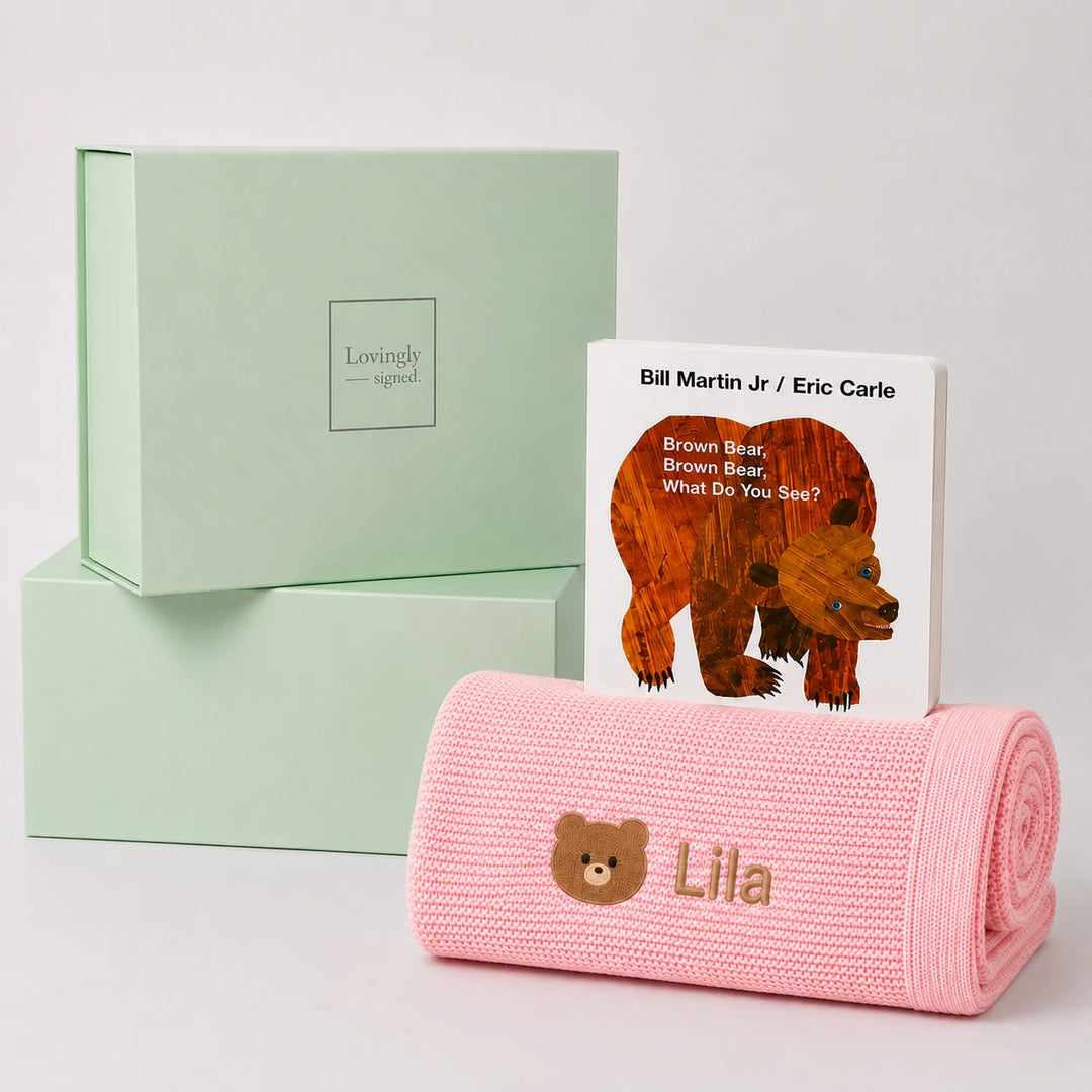 A personalized baby blanket in pink with an embroidered teddy bear face and the name 'Lila', displayed next to a children's book titled 'Brown Bear, Brown Bear: What Do You See?' by Bill Martin Jr. and Eric Carle, with two mint green 'Lovingly Signed' gift boxes in the background.