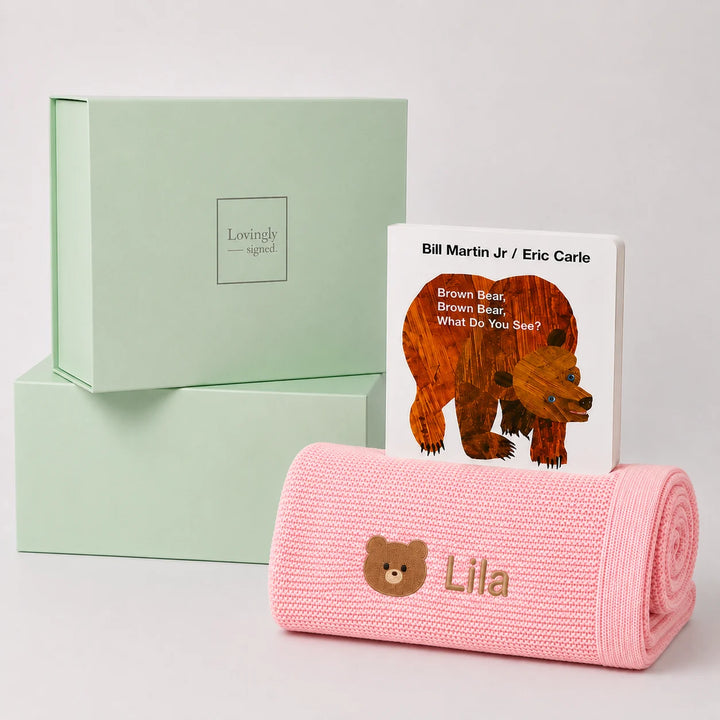 A personalized baby blanket in pink with an embroidered teddy bear face and the name 'Lila', displayed next to a children's book titled 'Brown Bear, Brown Bear: What Do You See?' by Bill Martin Jr. and Eric Carle, with two mint green 'Lovingly Signed' gift boxes in the background.