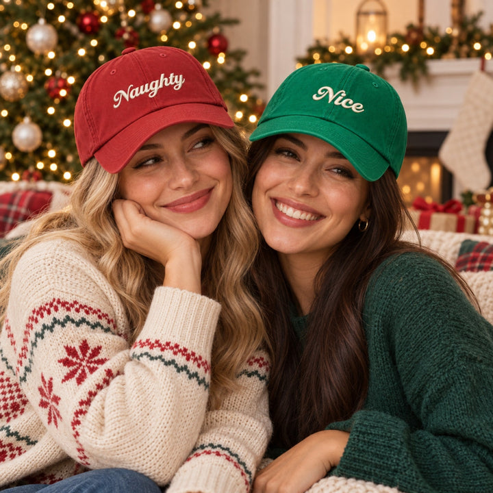 Two best friends wearing red “Naughty” and green “Nice” embroidered Christmas caps, smiling together in a cosy festive setting.