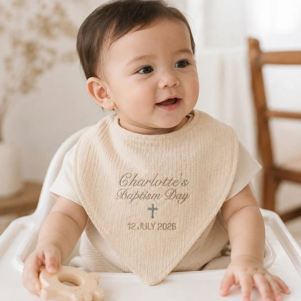 Baby wearing personalised beige baptism bib with embroidered name Charlotte, cross and baptism date