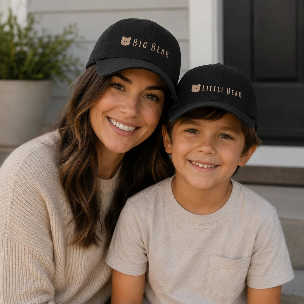 Woman and child wearing matching black hats with 'BIG BEAR' and 'LITTLE BEAR' text, sitting outdoors.