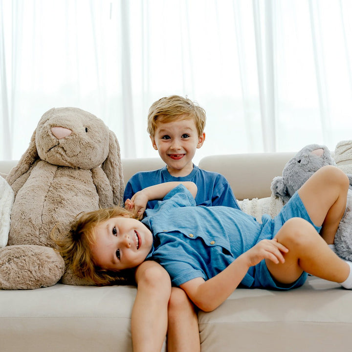 Two smiling kids sitting on a couch with Jellycat bunnies