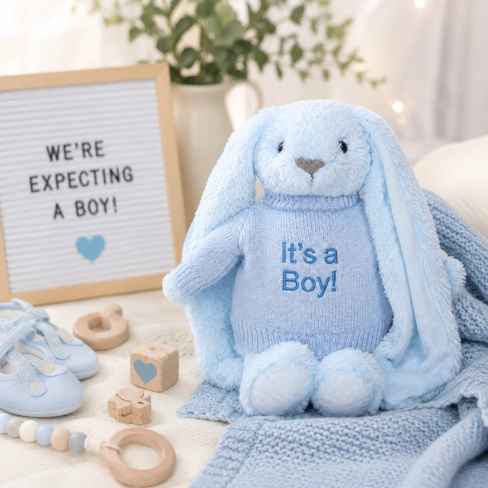 Blue bunny soft toy wearing an “It’s a Boy!” jumper, styled in a baby boy gender reveal setup with blue baby shoes, wooden toys, blanket, and a letter board.