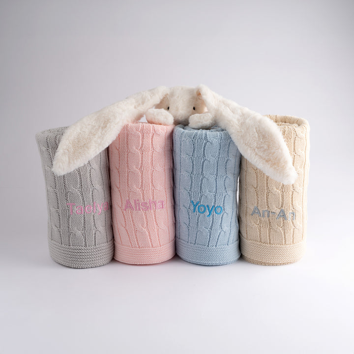 Soft and stylish cable knit baby blanket in pink, grey, cream, and blue. Perfect for cribs, prams, and car seats.