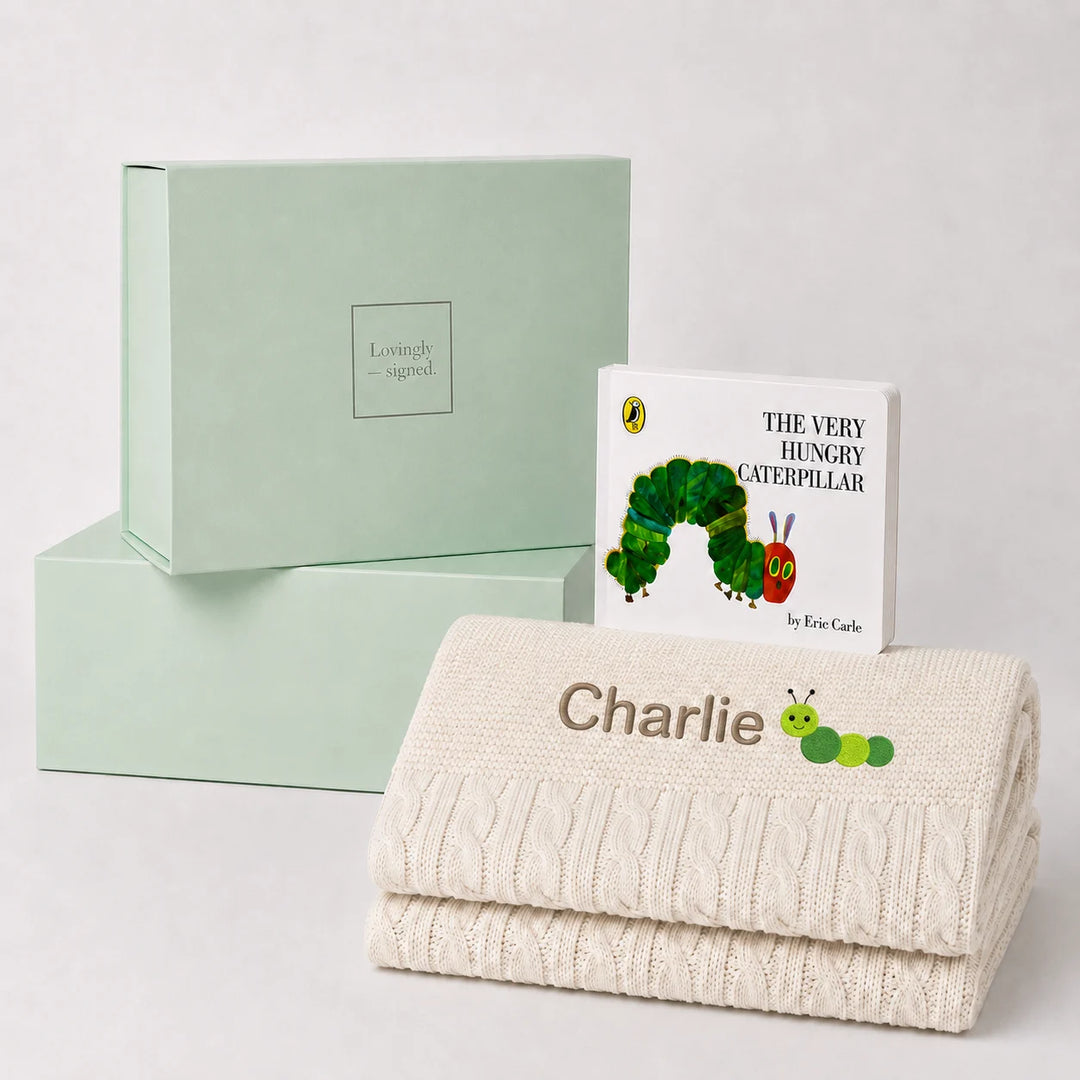 A personalized baby gift set featuring a cream cable-knit blanket with the name 'Charlie' and a caterpillar patch, a children's book of 'The Very Hungry Caterpillar' by Eric Carle, and a mint green 'Lovely Gift' box, arranged on a light gray background.