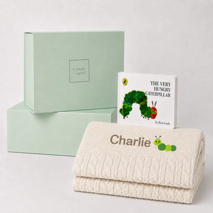 A personalized baby gift set featuring a cream cable-knit blanket with the name 'Charlie' and a caterpillar patch, a children's book of 'The Very Hungry Caterpillar' by Eric Carle, and a mint green 'Lovely Gift' box, arranged on a light gray background.