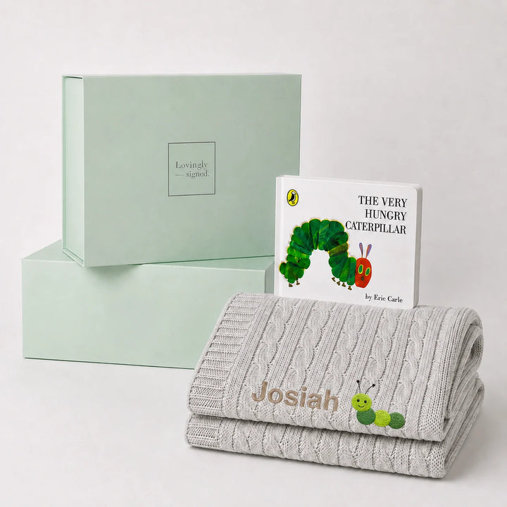 A personalized baby gift set featuring a light gray cable-knit blanket with the name 'Josiah' and a caterpillar patch, a children's book of 'The Very Hungry Caterpillar' by Eric Carle, and mint green gift boxes.