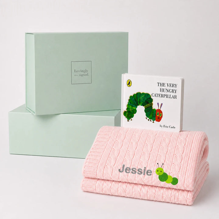 A personalized baby gift set featuring a pink cable-knit blanket with the name 'Jessie' and a caterpillar illustration, a children's book of 'The Very Hungry Caterpillar' by Eric Carle, and two mint green gift boxes.