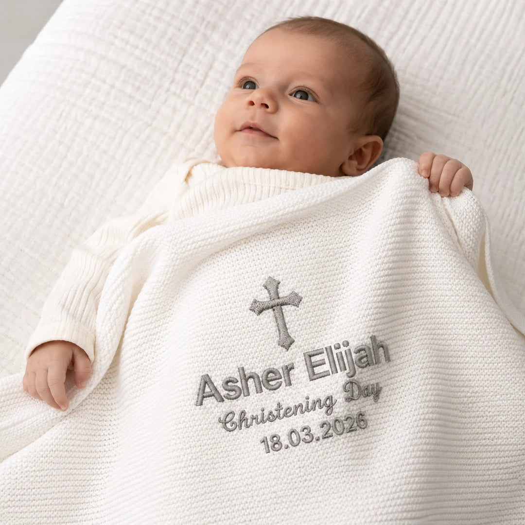 Baby wrapped in a white blanket with embroidered text on a neutral background