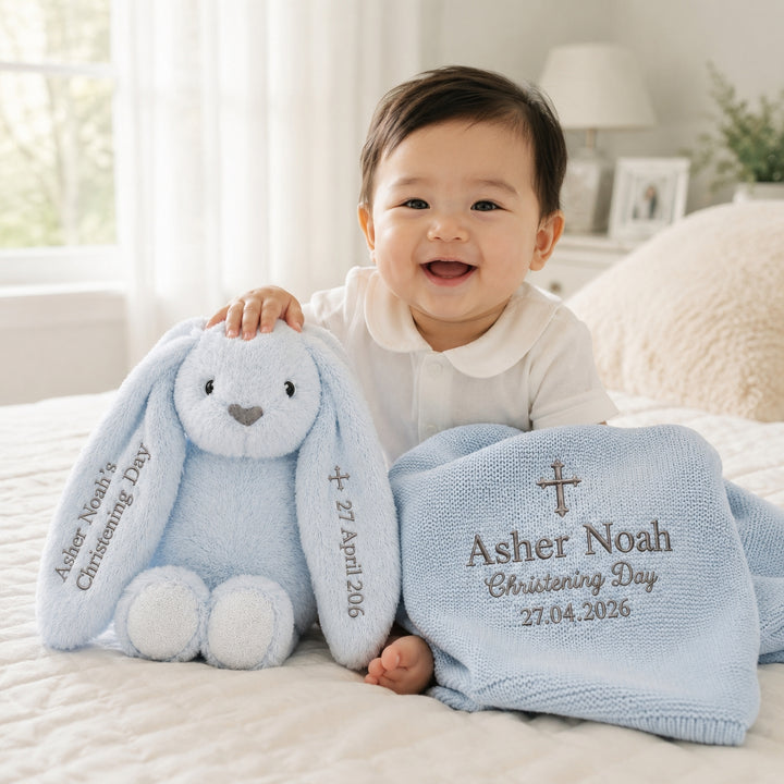 Smiling baby boy sitting on a bed with a personalised blue bunny soft toy and matching blue embroidered christening blanket, featuring the name “Asher Noah”, cross design, “Christening Day”, and date.