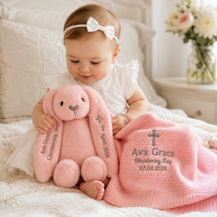Baby girl in a white dress holding a personalised pink bunny soft toy beside a matching pink embroidered christening blanket, featuring the name “Ava Grace”, cross design, “Christening Day”, and date.