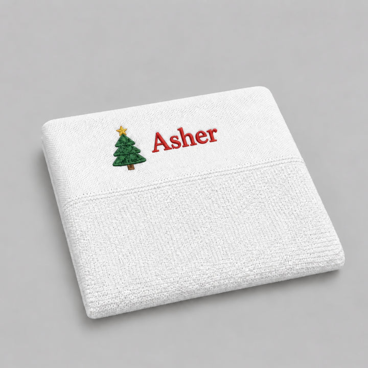 Folded white textured blanket embroidered with a green Christmas tree and the name “Asher” in red thread, photographed on a light grey studio background.