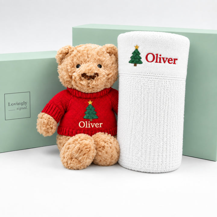 teddy bear wearing a red knitted Christmas jumper embroidered with a Christmas tree and the name “Oliver,” displayed beside a rolled white personalised blanket with matching Christmas tree embroidery and the name “Oliver,” against mint green Lovingly Signed gift boxes.