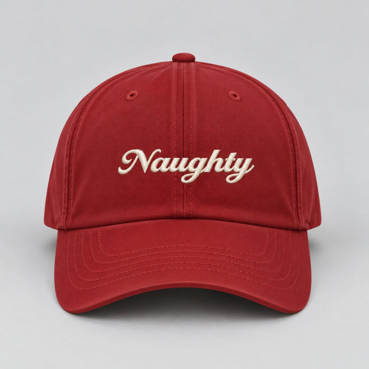 Close-up front view of a red Christmas baseball cap featuring “Naughty” embroidered in cream cursive lettering.
