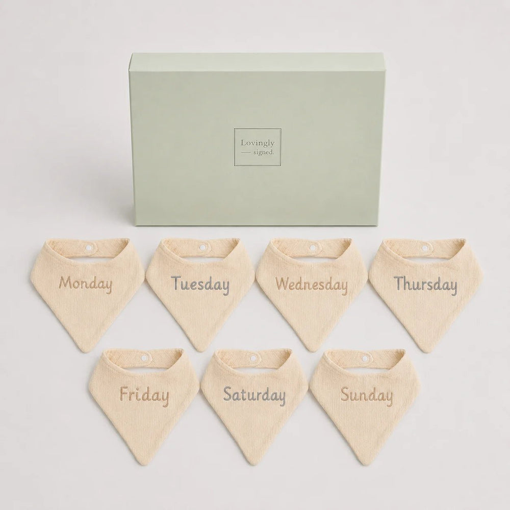 Set of seven beige bandana bibs labeled for each day of the week in front of a green box with 'Lovingly Signed' branding.