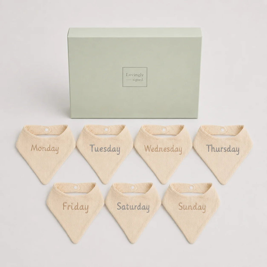 Set of seven beige bandana bibs labeled for each day of the week in front of a green box with 'Lovingly Signed' branding.