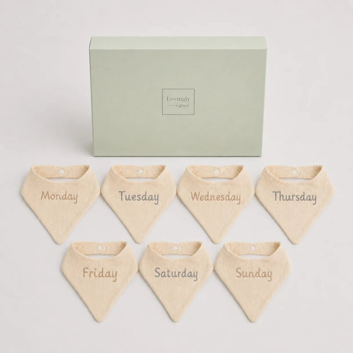 Set of seven beige bandana bibs labeled for each day of the week in front of a green box with 'Lovingly Signed' branding.
