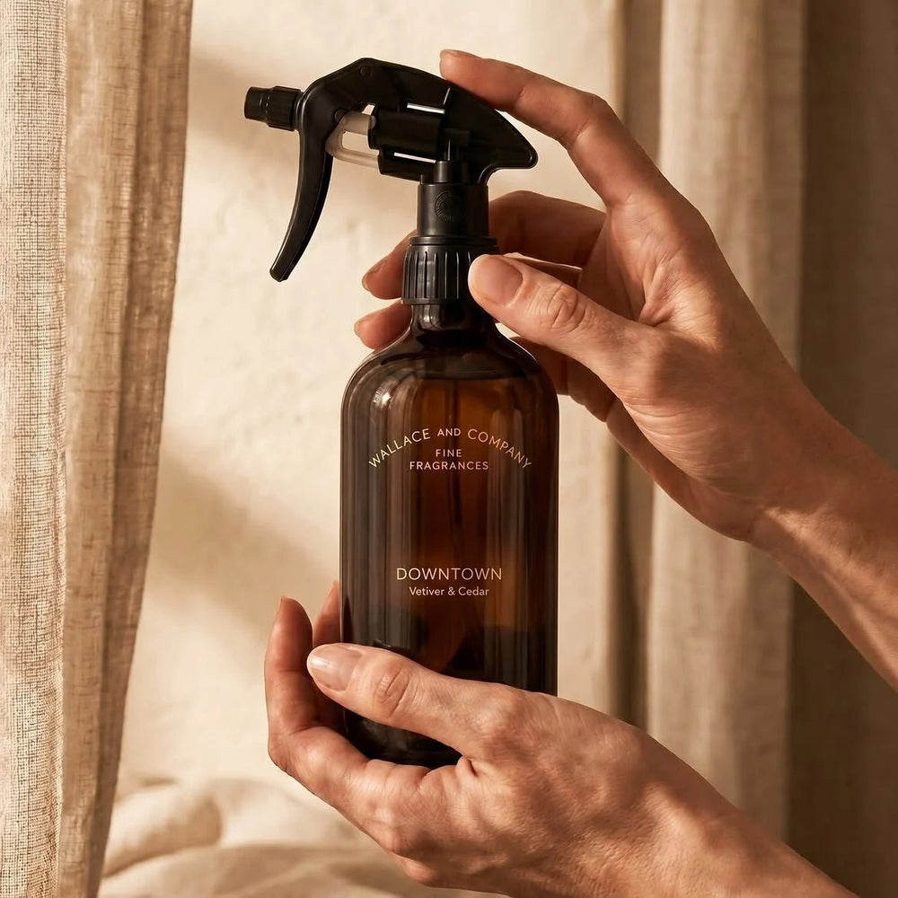 A person holds a dark amber glass spray bottle with a black trigger sprayer. The bottle is labelled 'WALLACE AND COMPANY FINE FRAGRANCES' and 'DOWNTOWN Vetiver & Cedar'.