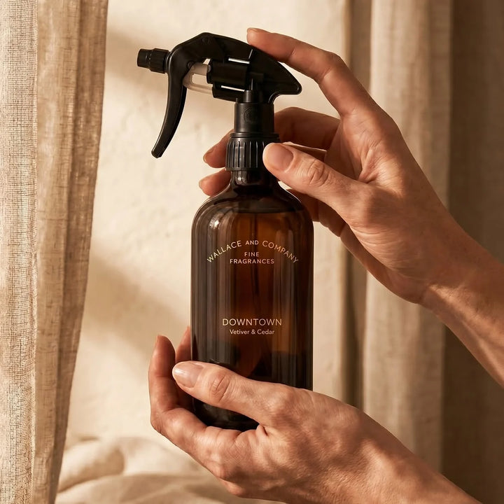 A person holds a dark amber glass spray bottle with a black trigger sprayer. The bottle is labelled 'WALLACE AND COMPANY FINE FRAGRANCES' and 'DOWNTOWN Vetiver & Cedar'.