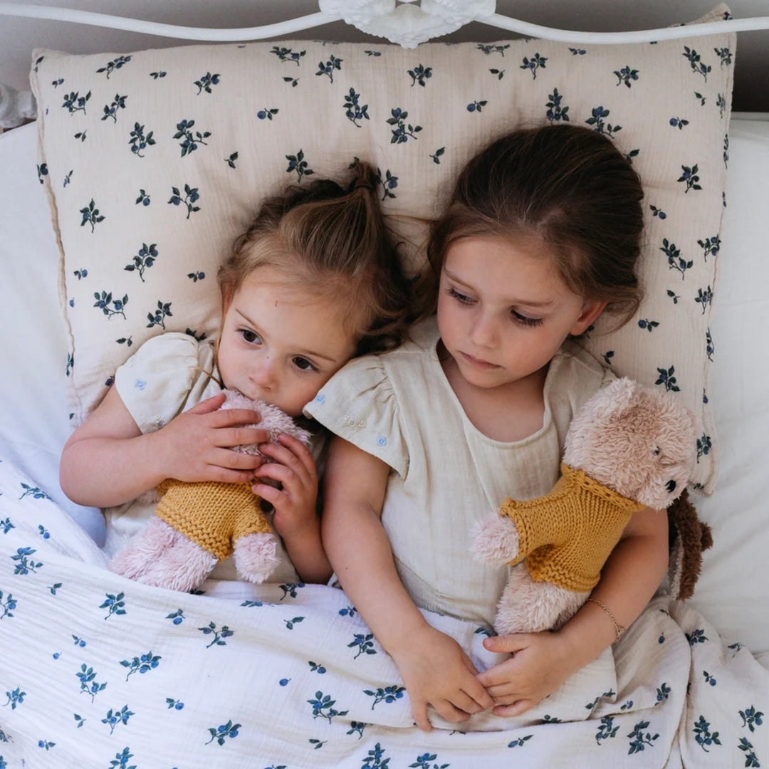Two young girls relaxing in bed with floral bedding, each cuddling a soft teddy bear dressed in a mustard yellow knitted jumper.