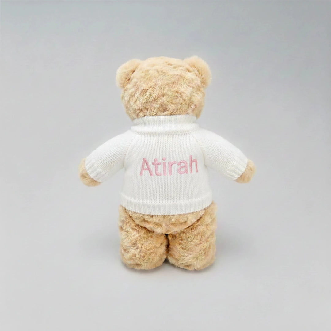 Beige teddy bear wearing a white sweater with 'Atirah' embroidered on a gray background