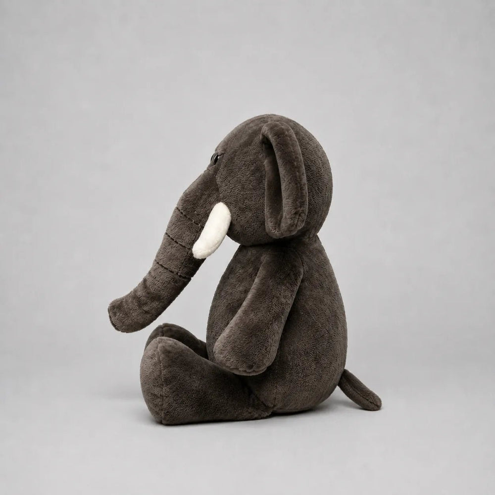 A dark grey plush elephant toy sitting down, featuring white tusks and a soft textured fabric, photographed against a neutral gray background.