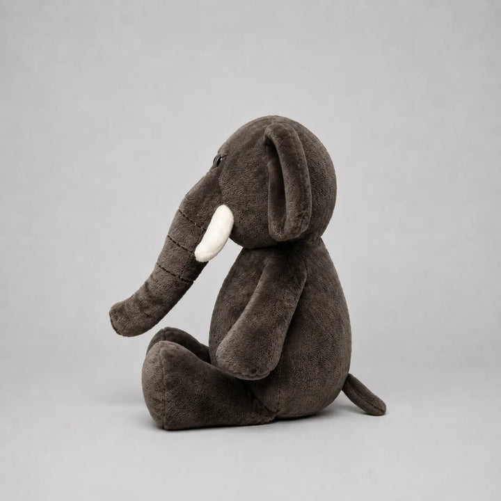 A dark grey plush elephant toy sitting down, featuring white tusks and a soft textured fabric, photographed against a neutral gray background.