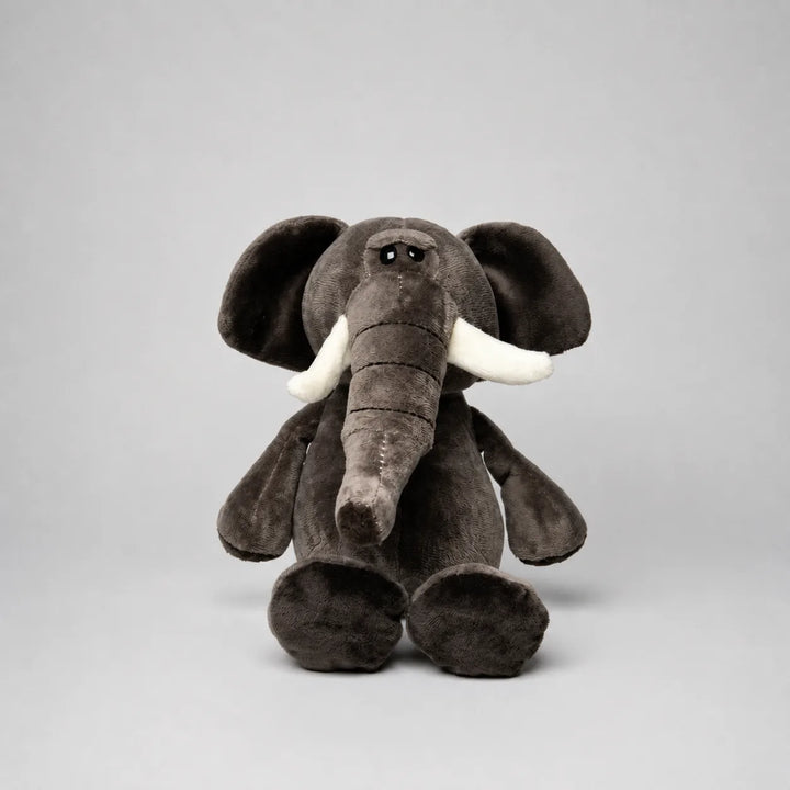 A plush stuffed elephant toy in dark brownish-gray with white tusks, photographed against a plain light gray studio background.