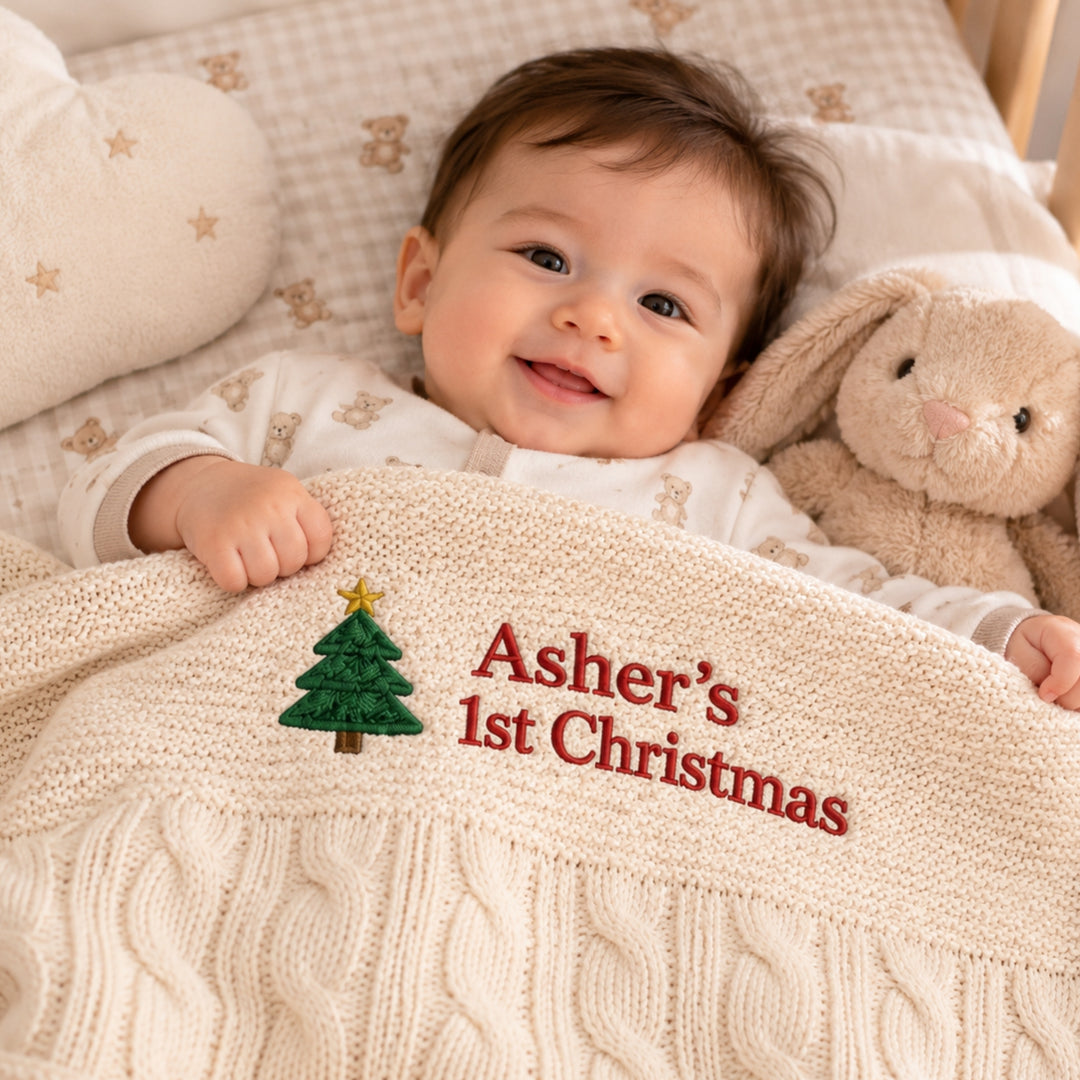 Smiling baby boy lying in a cosy cot, covered with a personalised cream cable-knit blanket embroidered with a Christmas tree and “Asher’s 1st Christmas.”