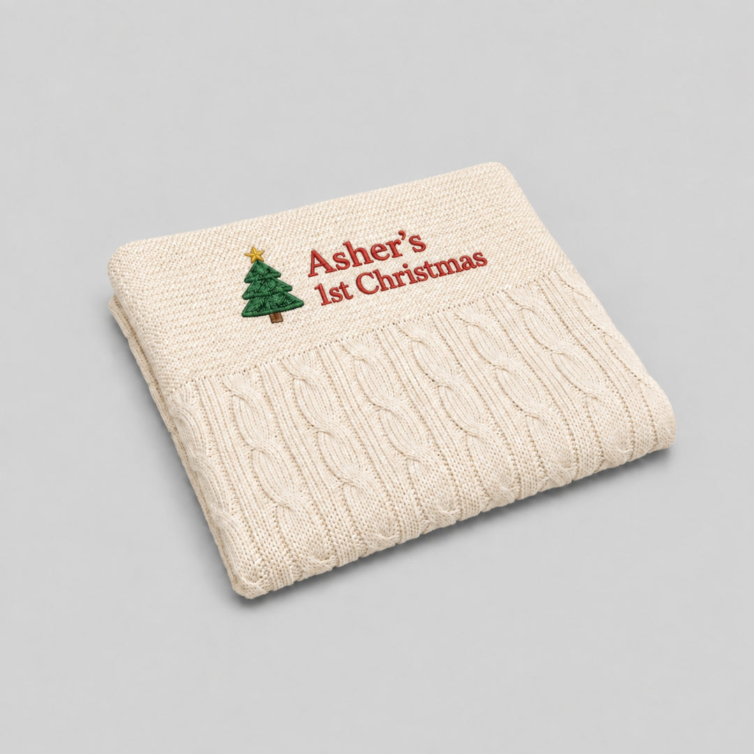 Cream cable-knit baby blanket personalised with a Christmas tree motif and “Asher’s 1st Christmas” embroidered in red.
