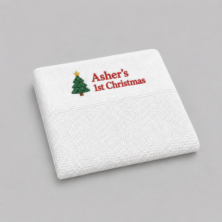 White personalised knitted Christmas baby blanket embroidered with a Christmas tree and “Asher’s 1st Christmas” in red, photographed on a light grey studio background.