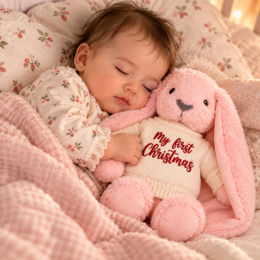 Sleeping baby cuddled beside a pink plush bunny wearing a cream knitted jumper embroidered with “My First Christmas” in red.