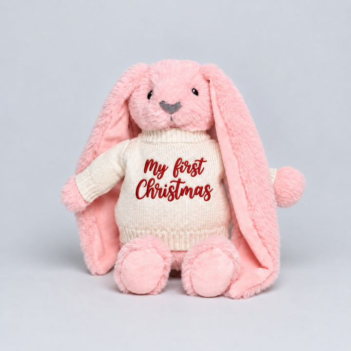 Pink plush bunny with long floppy ears wearing a cream knitted jumper embroidered with “My First Christmas” in red.