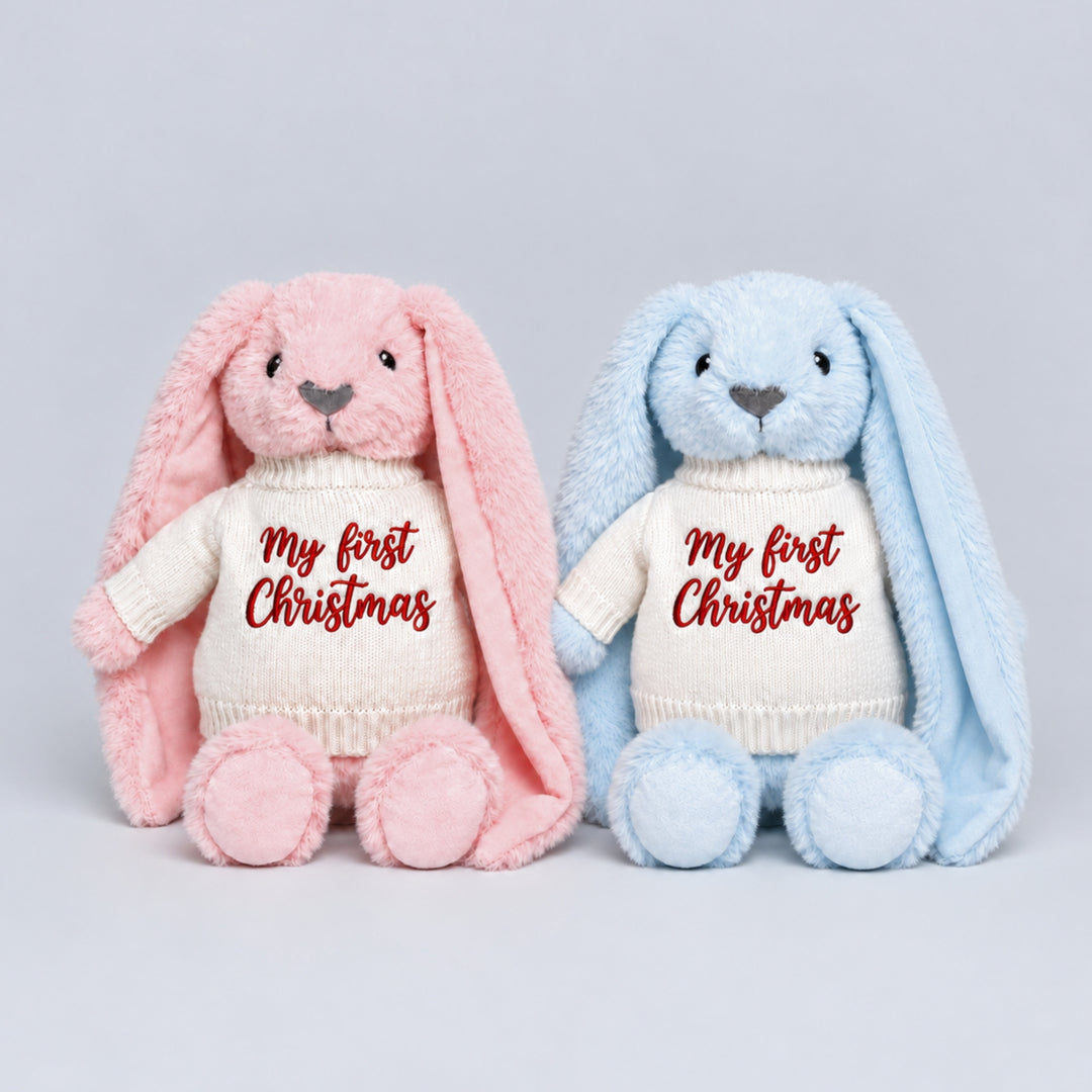 Pink and blue plush bunnies sitting side by side, wearing matching cream knitted jumpers embroidered with “My First Christmas” in red.