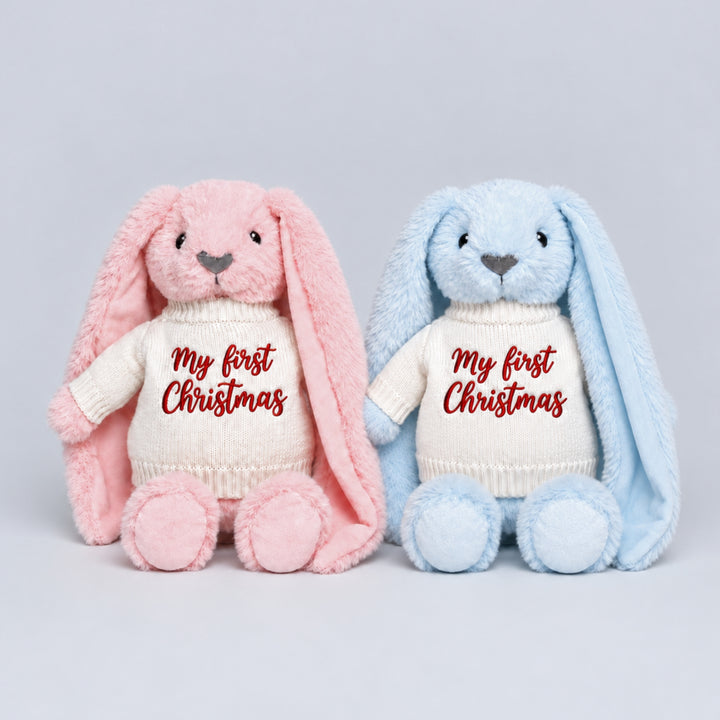 Pink and blue plush bunnies sitting side by side, wearing matching cream knitted jumpers embroidered with “My First Christmas” in red.
