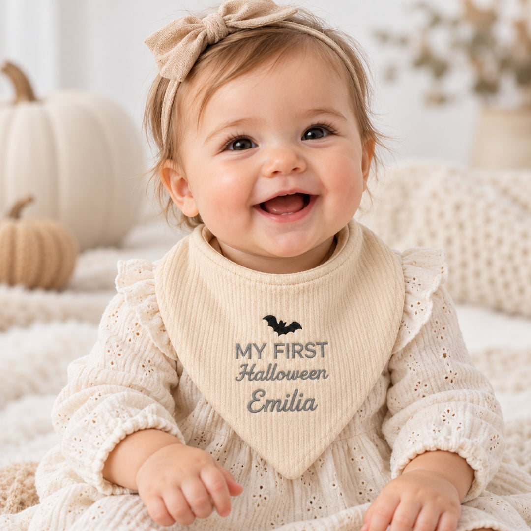 miling baby girl wearing a personalised cream “My First Halloween” bandana bib embroidered with a black bat and the name Emilia, dressed in a cream outfit with a matching bow headband.