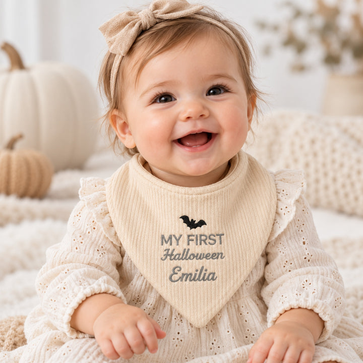 miling baby girl wearing a personalised cream “My First Halloween” bandana bib embroidered with a black bat and the name Emilia, dressed in a cream outfit with a matching bow headband.