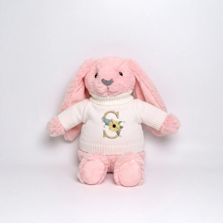 Pink plush bunny toy wearing a cream sweater with a letter 'S' and flower design on a light gray background